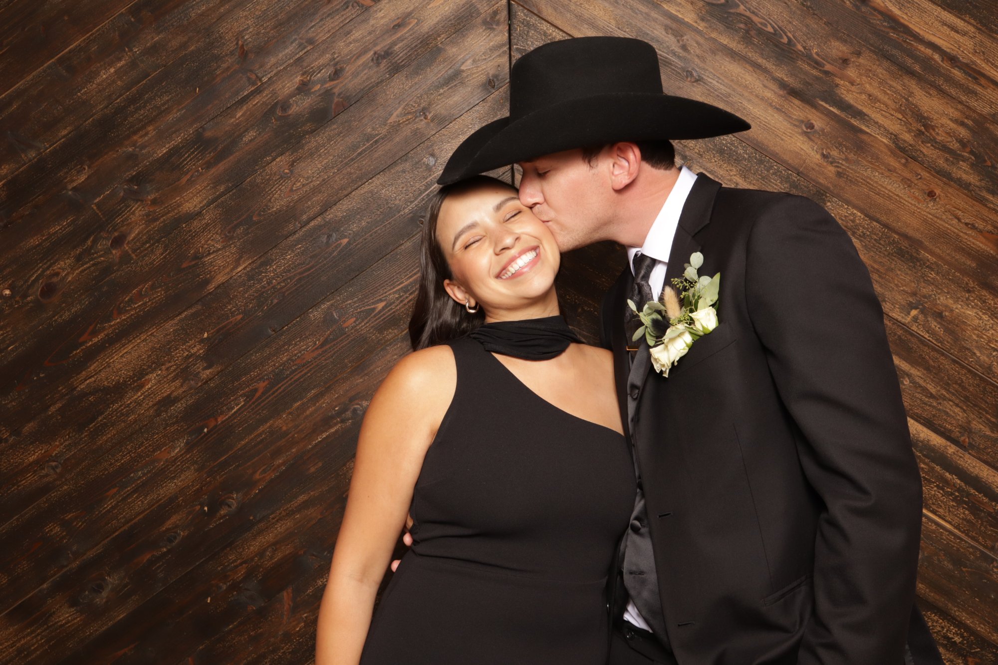 Man in a black cowboy hat and suit kissing a smiling woman's cheek against a dark wood backdrop