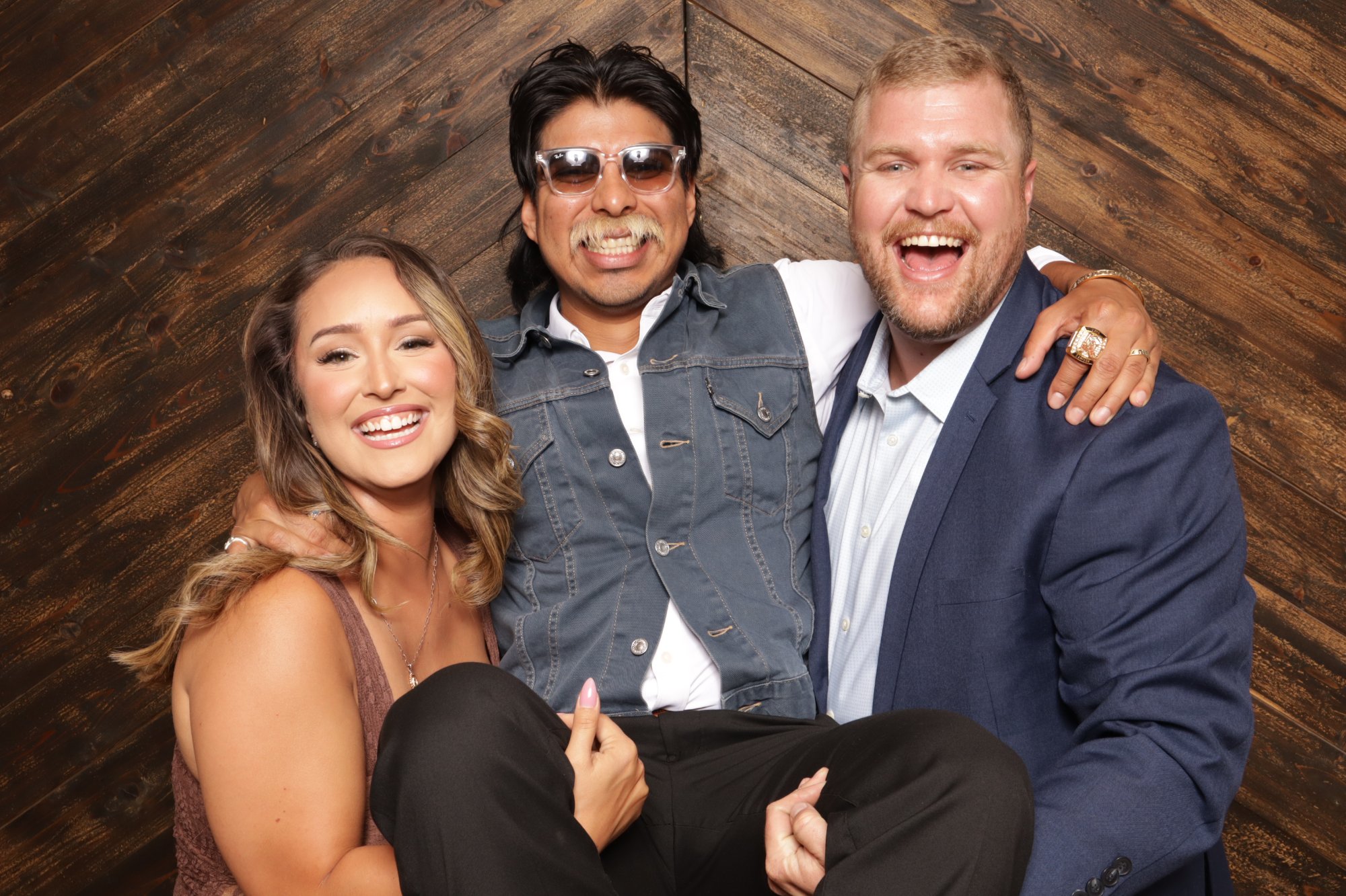 A woman and a man in a blue blazer carry a grinning friend in sunglasses and a denim vest against a dark wood backdrop
