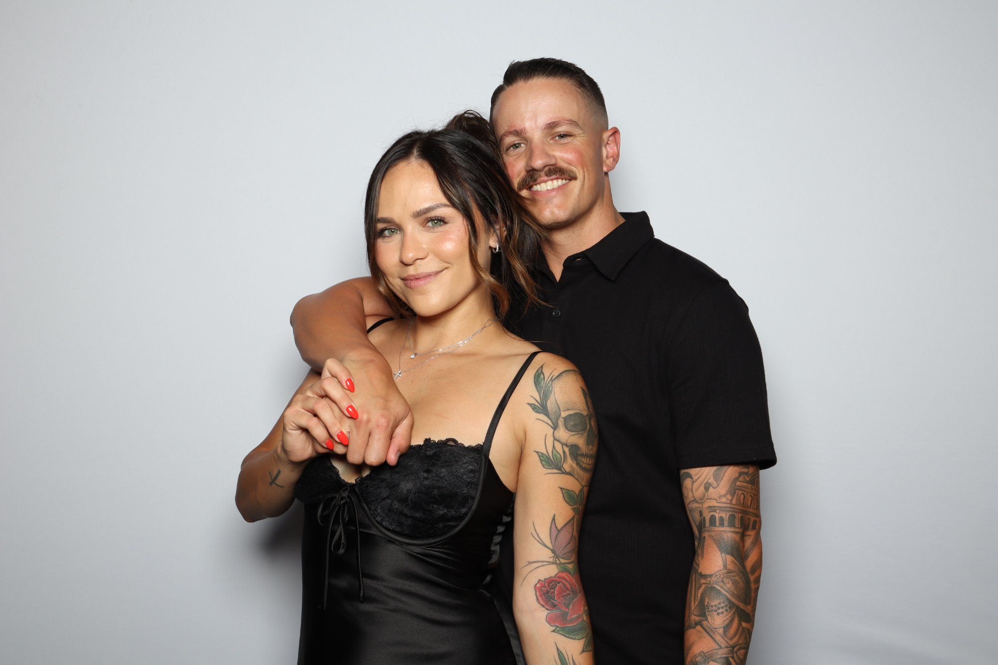 A couple at a light gray backdrop, he in a black shirt with his arm around her, she in a black satin slip dress