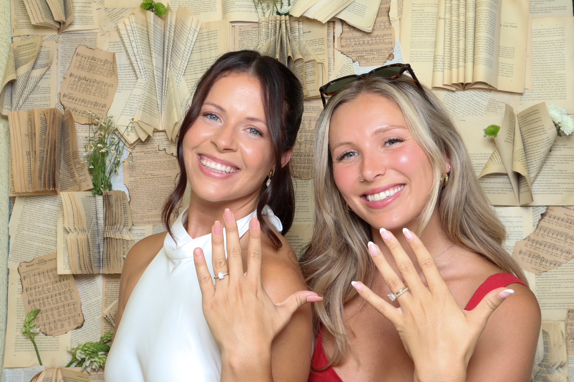 Two women showing their rings in front of a backdrop of folded book pages and sheet music