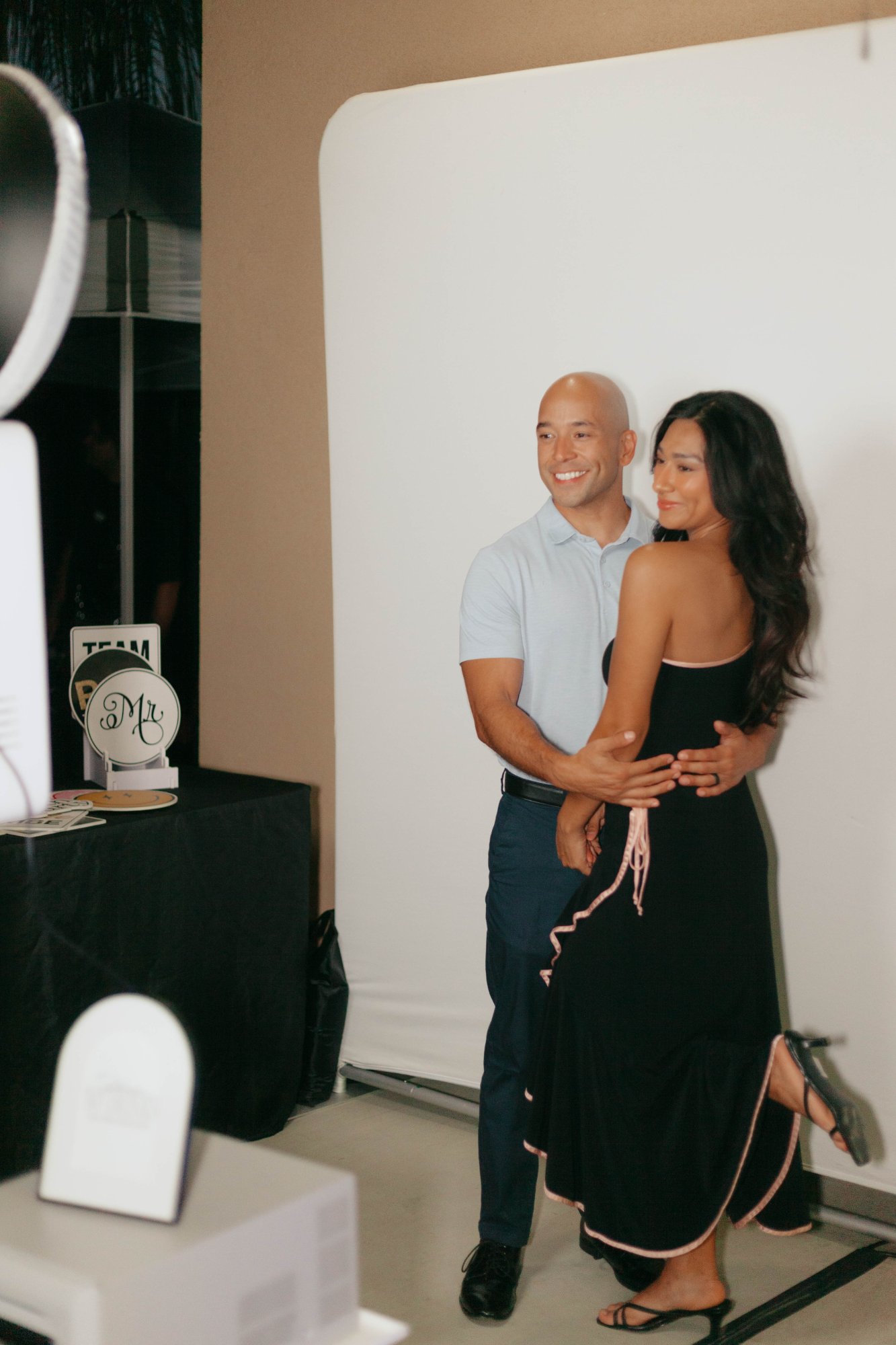 Couple embracing at a white backdrop, with a white photo booth and a prop table at left