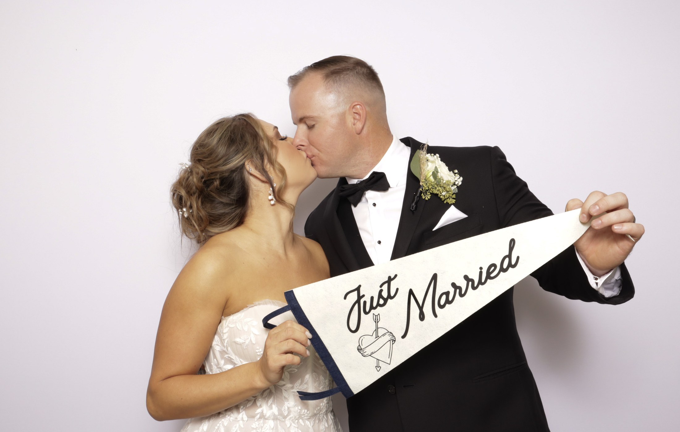 A bride in a strapless lace gown kisses a groom in a tuxedo as they hold a Just Married pennant before a white backdrop